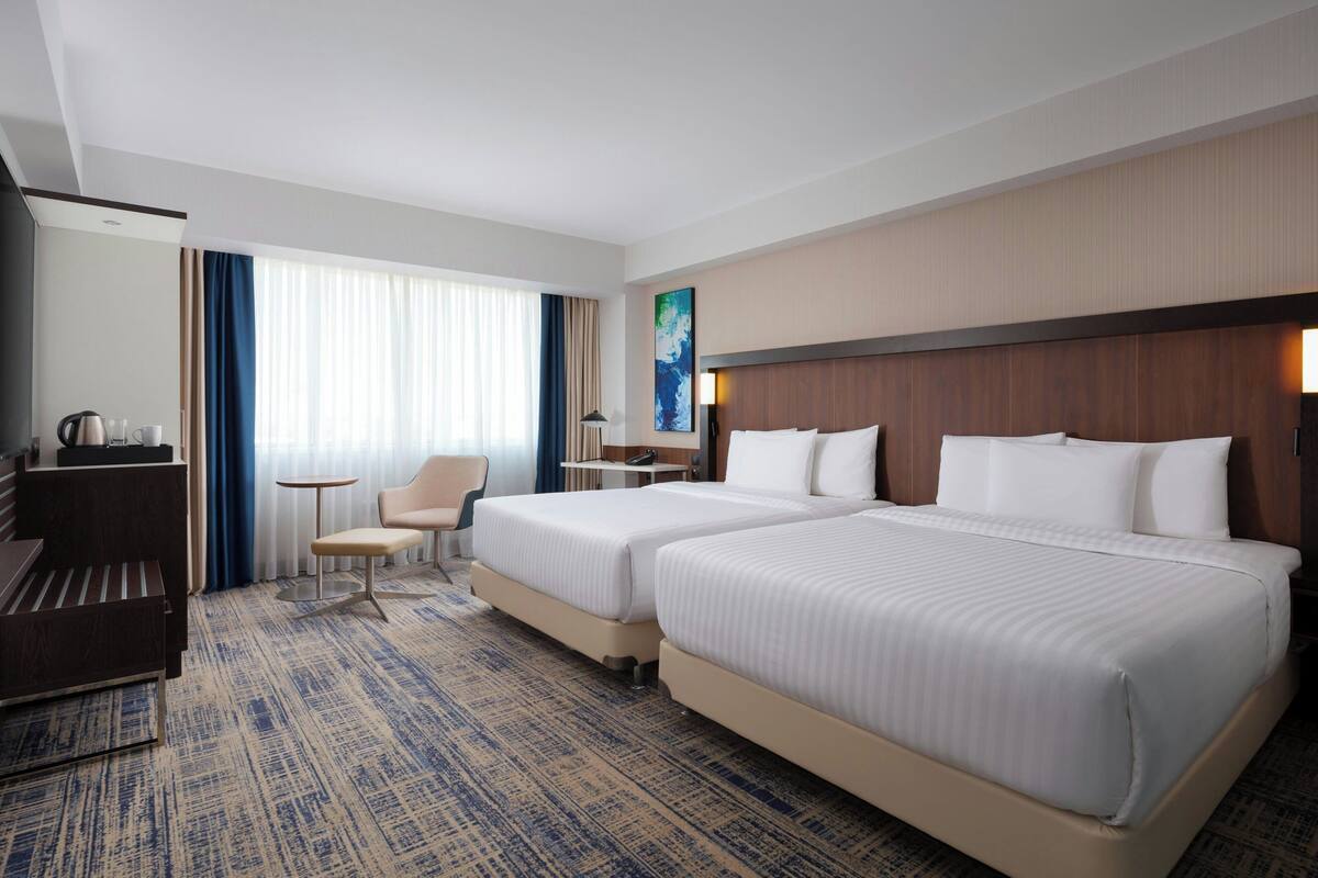 A hotel room with two beds, a desk, a chair, a lamp, and a painting on the wall.