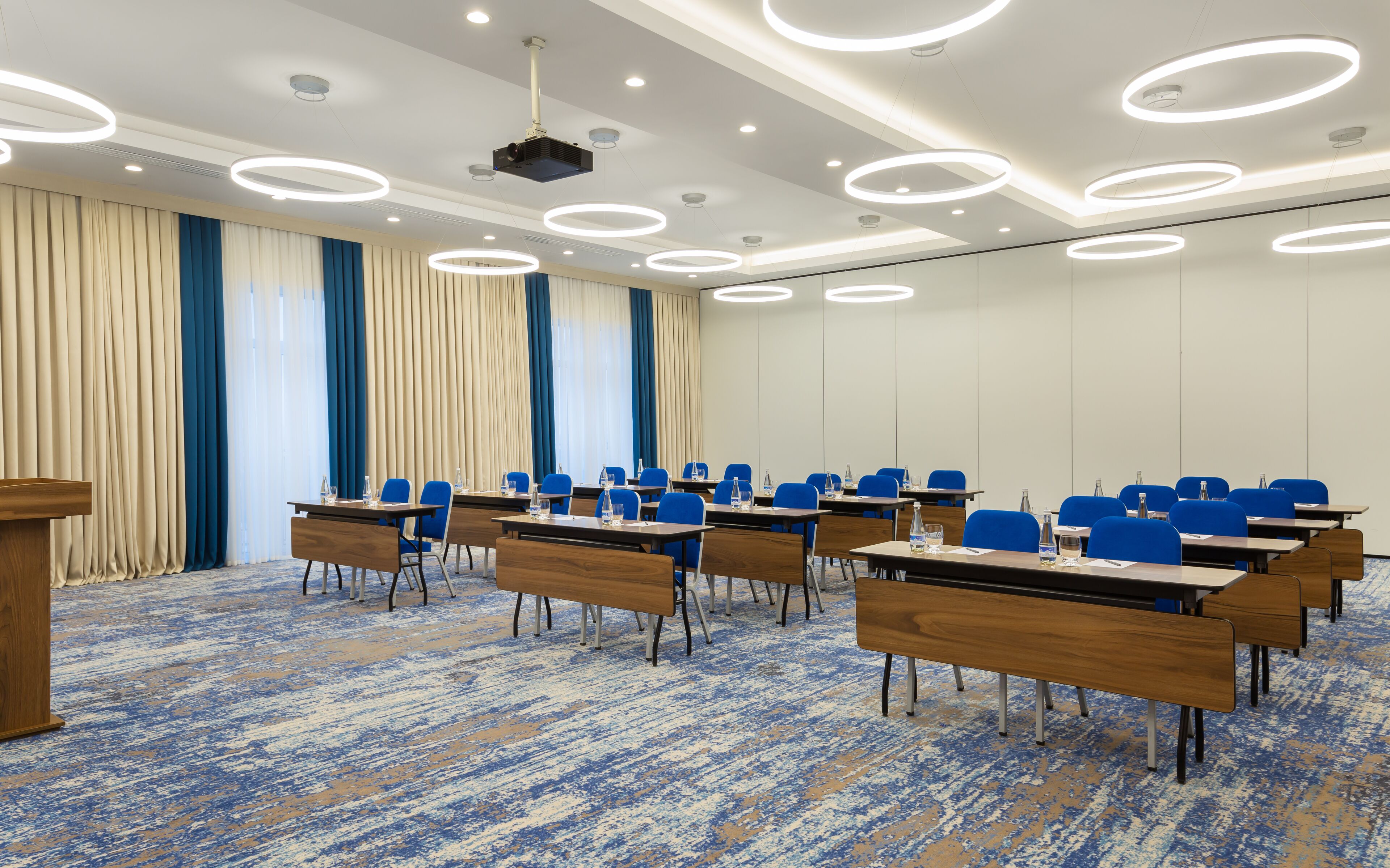 Un salón de conferencias con proyector, filas de sillas azules, mesas de madera y una alfombra con diseño azul y blanco.