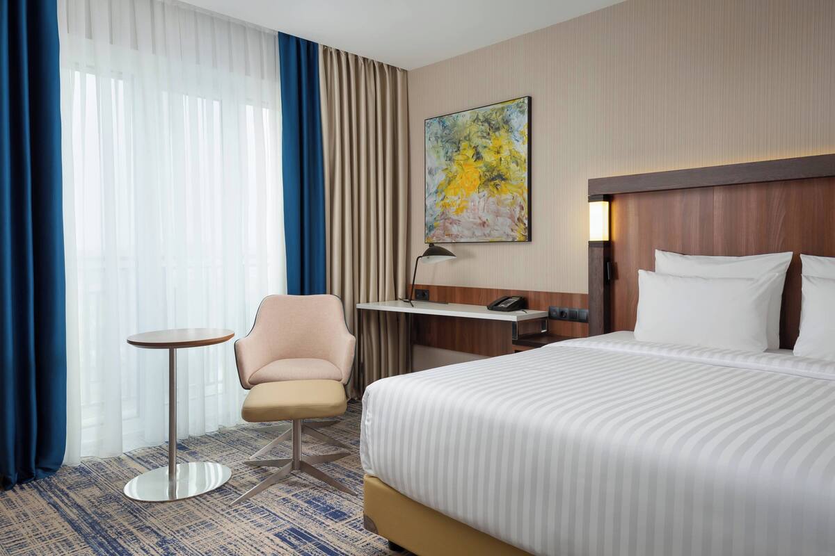 A hotel room with a bed, a desk, a chair, a small table, and a painting on the wall.
