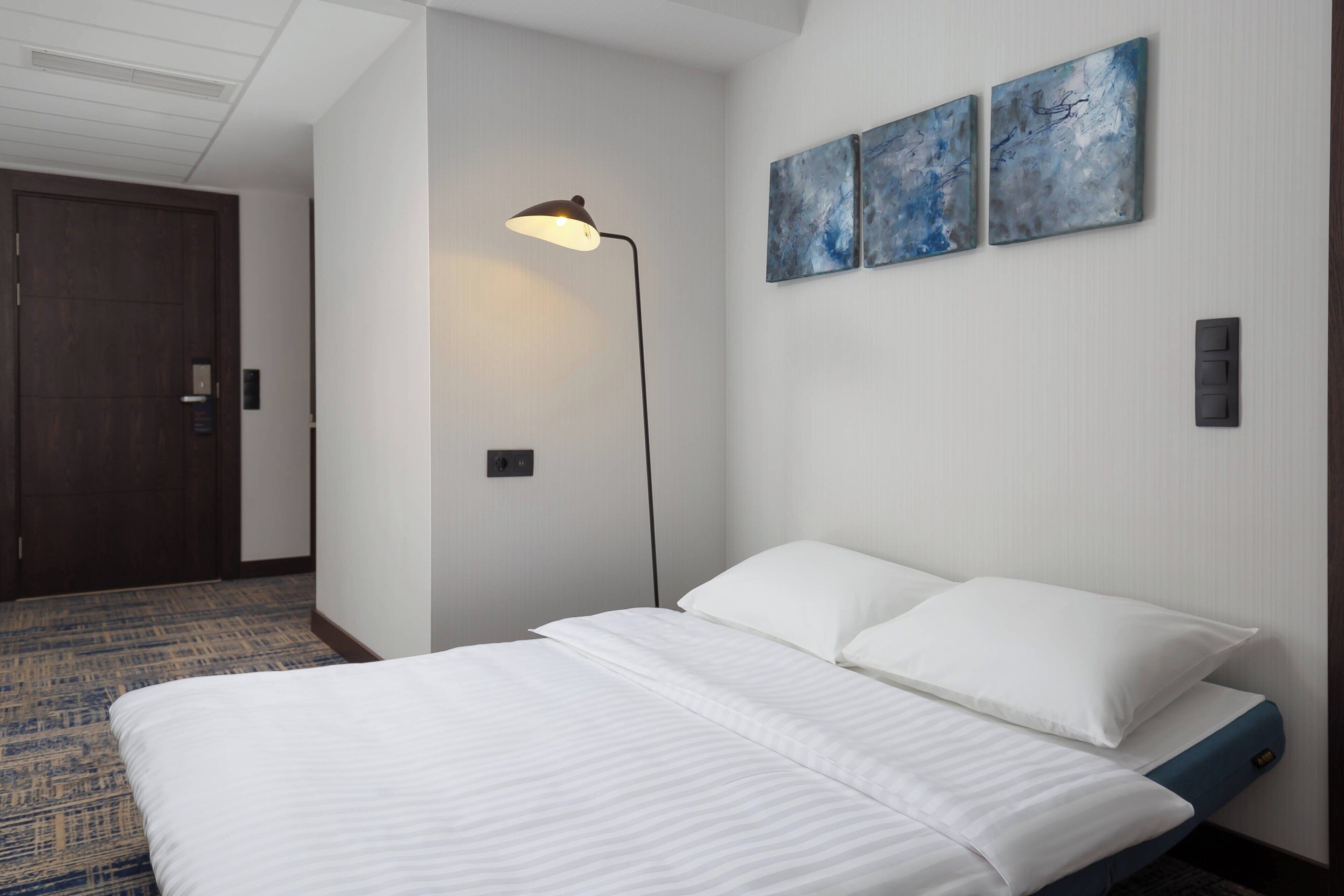 A hotel room with a bed, a floor lamp, and two abstract paintings on the wall.