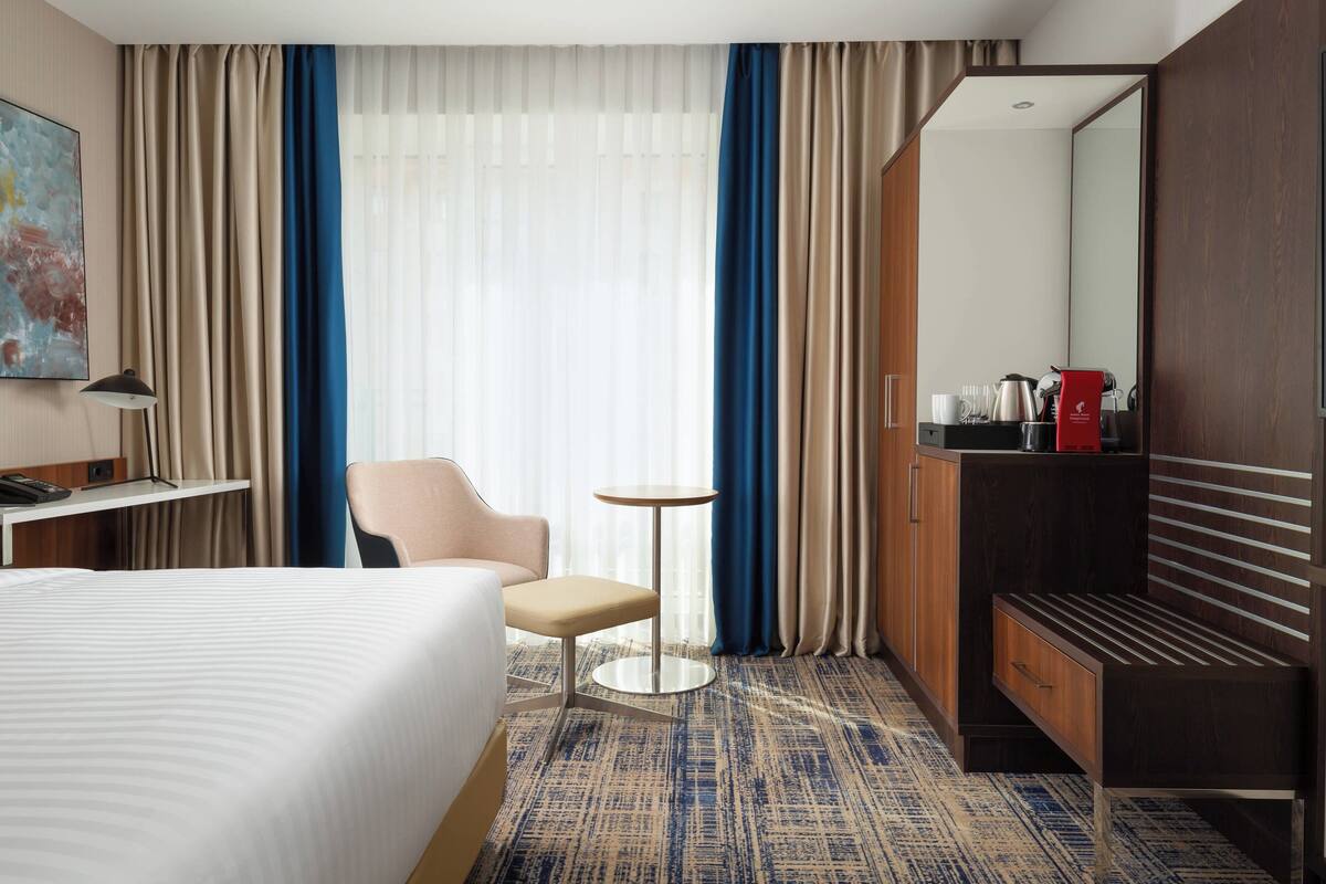 A hotel room with a bed, a desk, a chair, a small table, a wardrobe, and a large mirror.