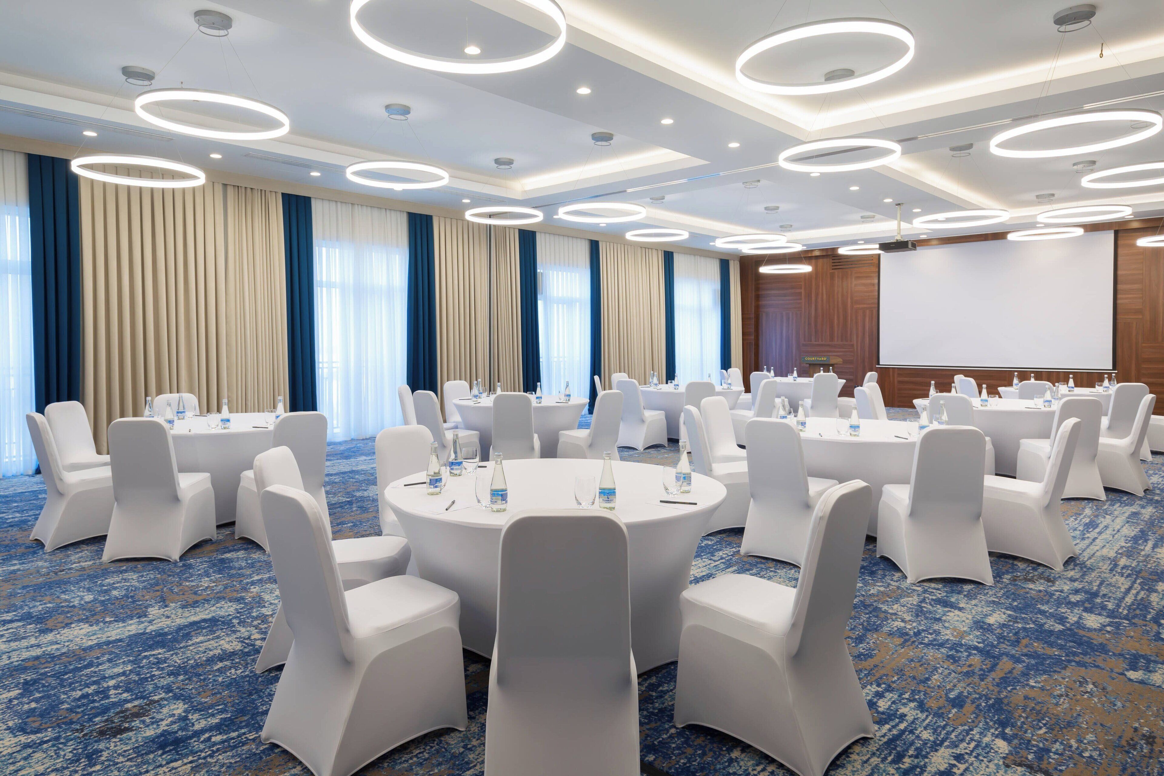 Une salle de conférence avec des tables rondes, des chaises blanches et un grand écran.