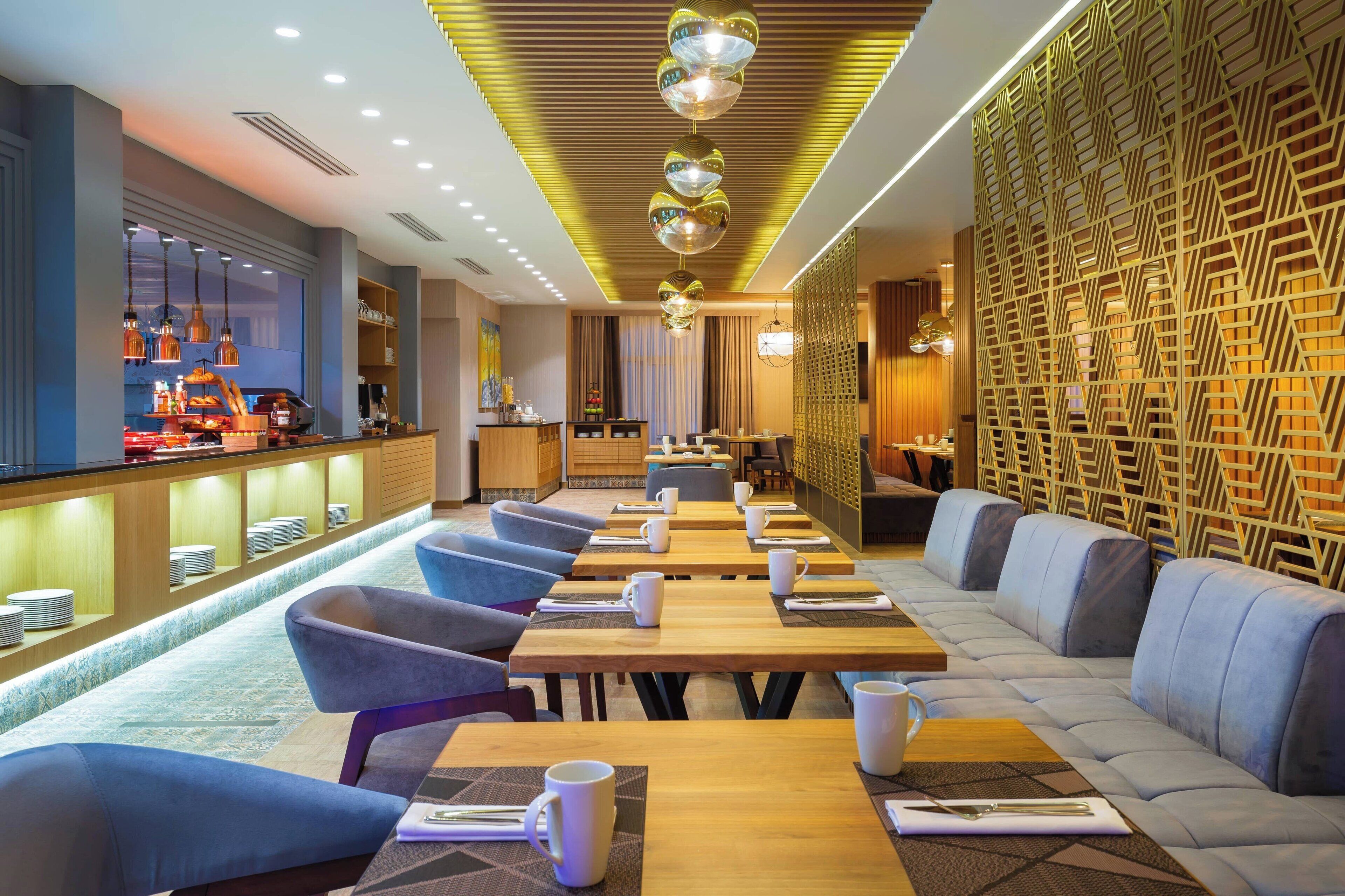 A modern restaurant with long wooden tables, blue cushioned seating, and a patterned wall.