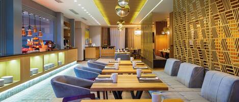 A modern restaurant with long wooden tables, blue cushioned seating, and a patterned wall.