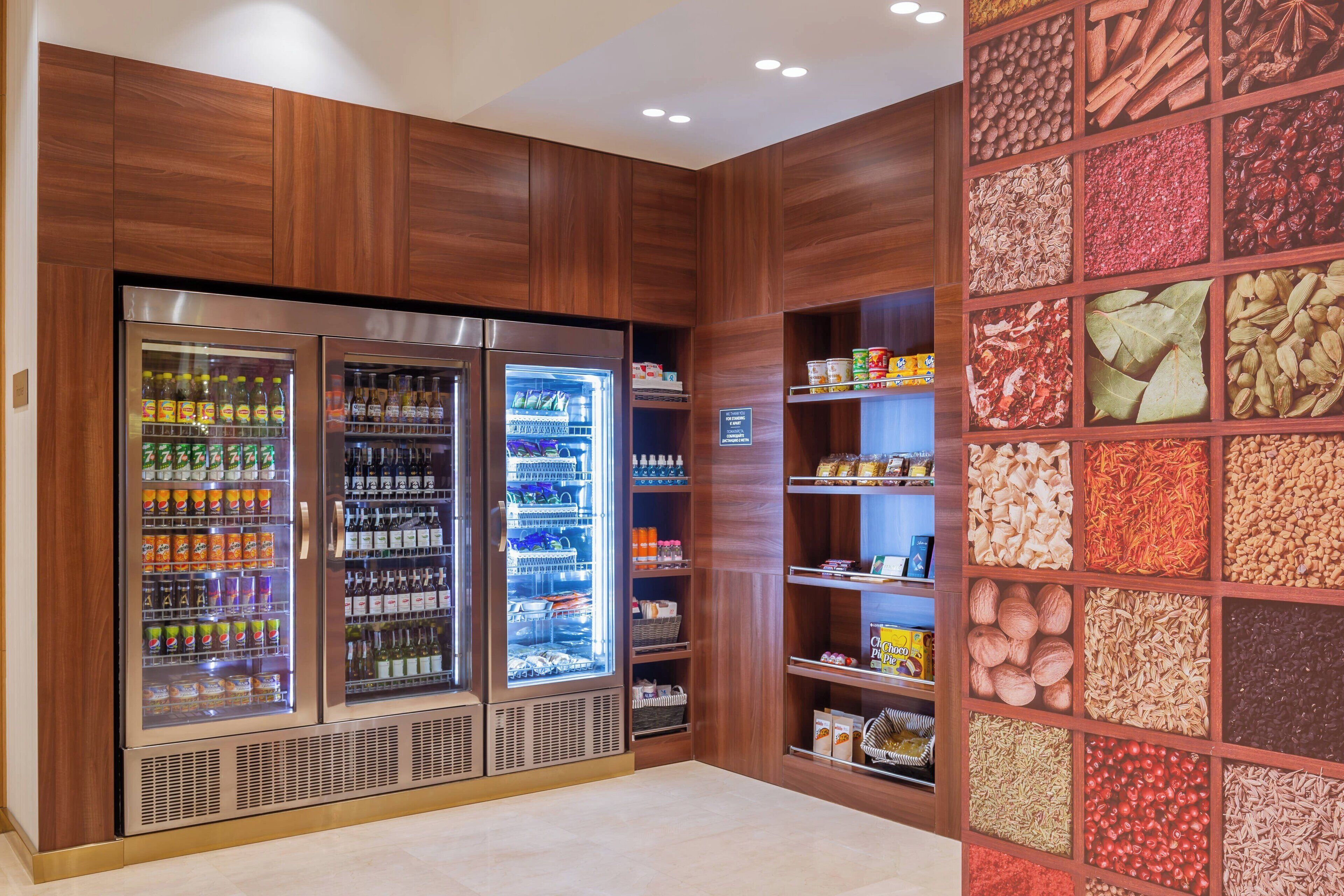 A modern hotel lobby with two large refrigerated beverage dispensers, a variety of snacks and condiments on shelves, and a wall display of spices and herbs.