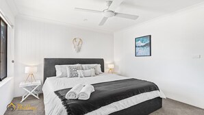 Un dormitorio con una cama, dos mesitas de noche, un ventilador de techo, una obra de arte en la pared y una ventana.