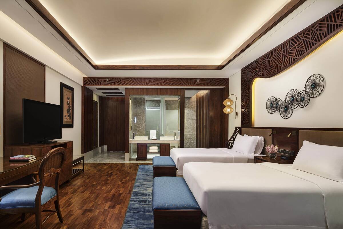A hotel room with a TV, a desk with books, two beds, a bathroom with a mirror, and a decorative wall piece.