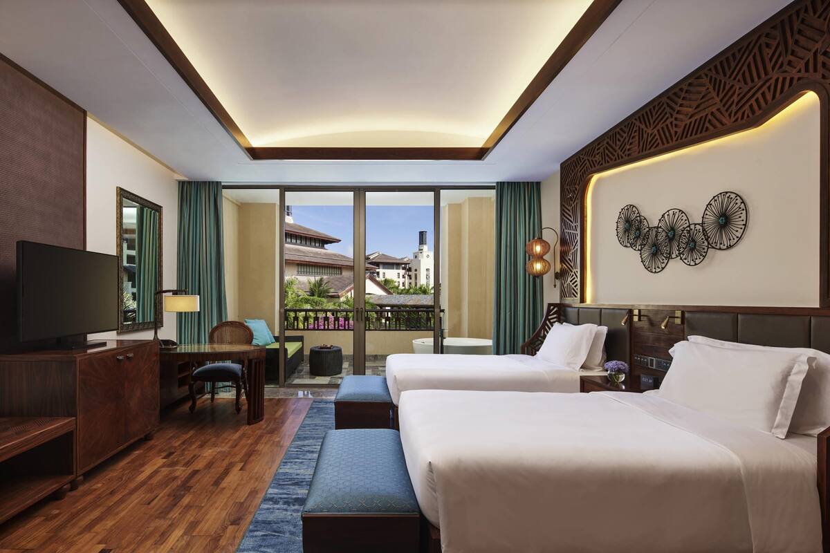 A hotel room with two beds, a TV, a desk, and a balcony with a view of buildings and palm trees.