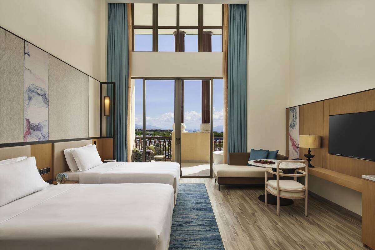 A hotel room with two beds, a balcony with a view, a TV, and a small table with a lamp.