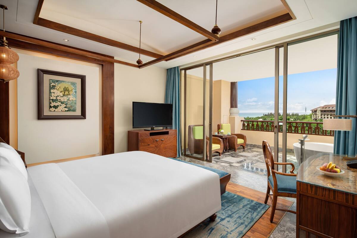 A hotel room with a large bed, a TV, a desk with a fruit bowl, and a balcony with a view of buildings and greenery.