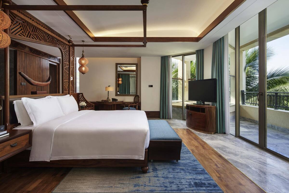 A spacious bedroom with a large bed, wooden furniture, a television, and a balcony with a view of palm trees.