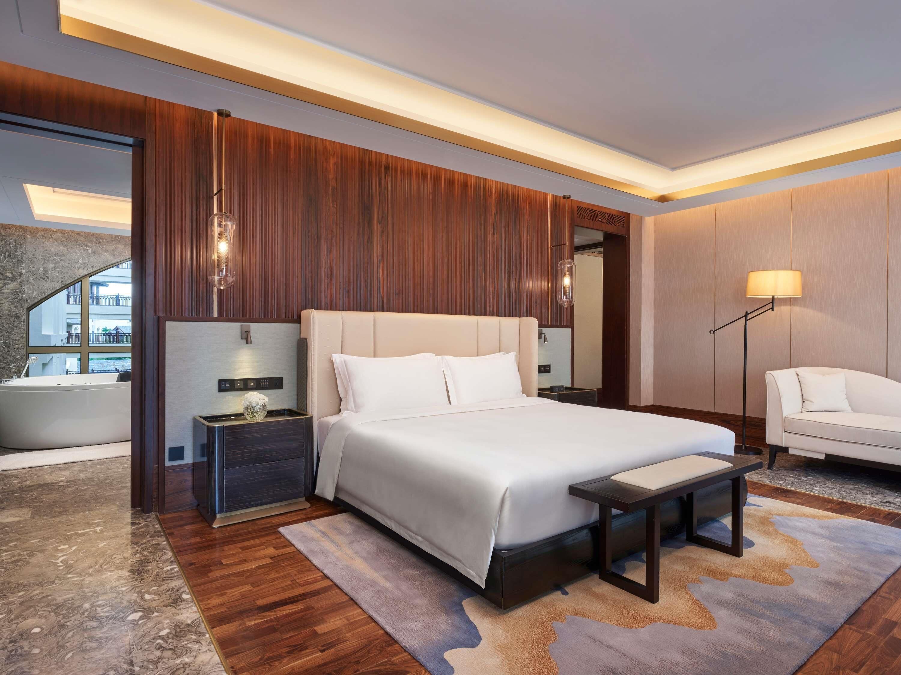 A modern hotel room with a large bed, a nightstand, a bedside table, a chair, a floor lamp, and a view of a building outside.