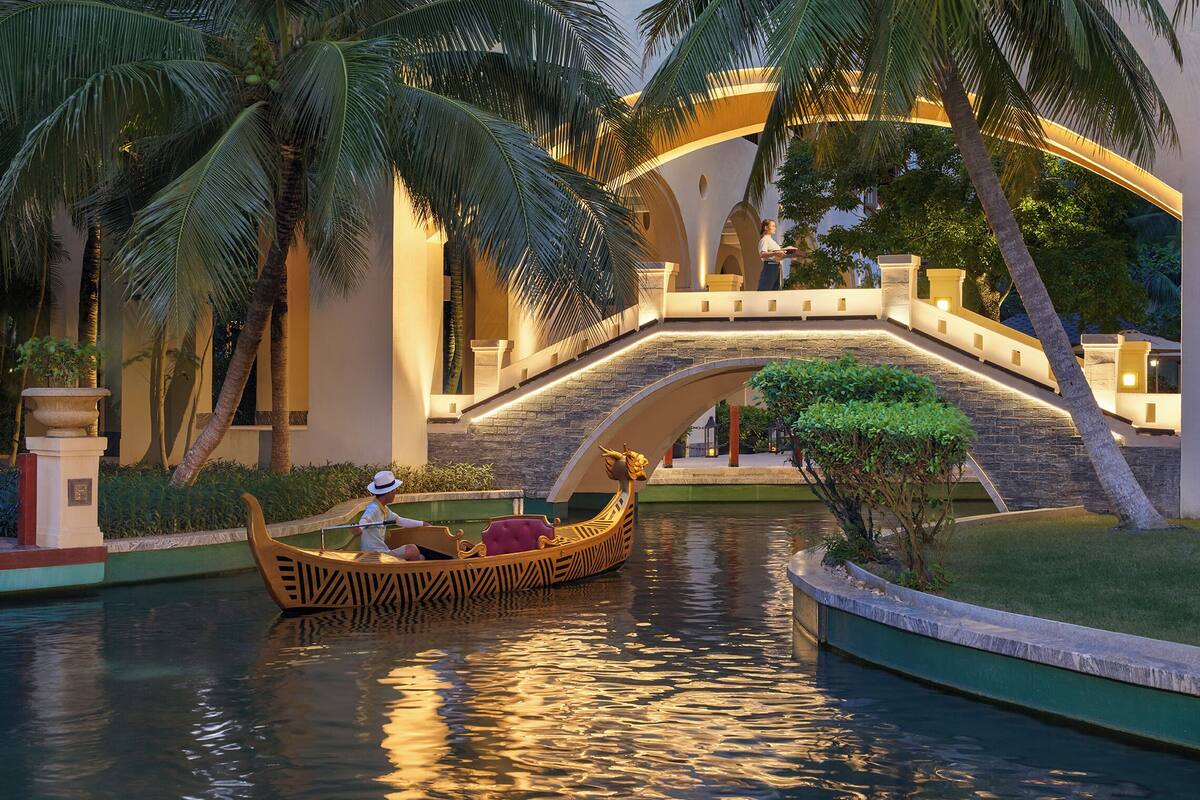 A canal with a traditional boat, palm trees, and a bridge with people walking.