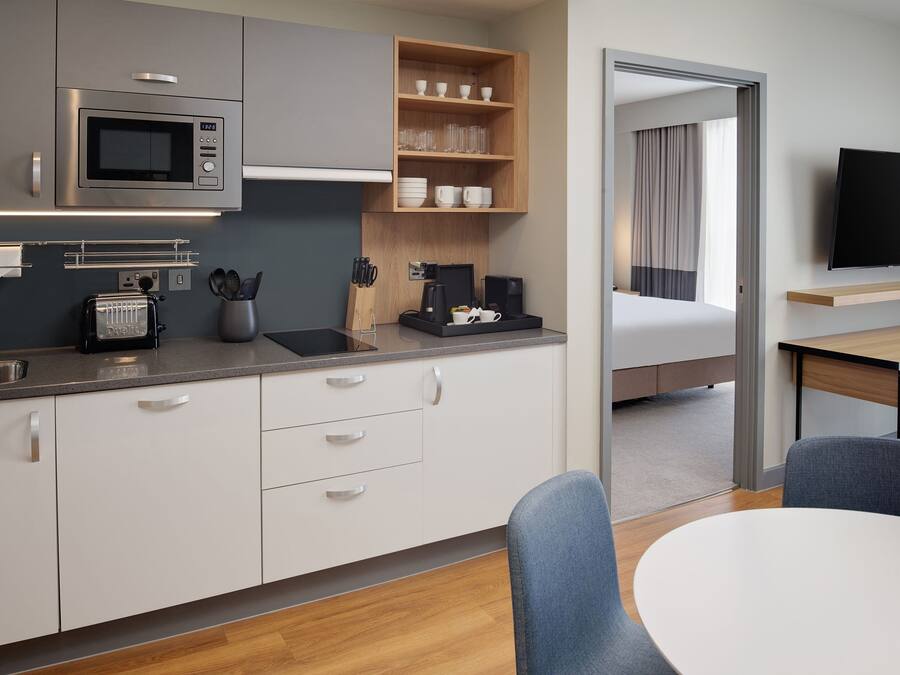 Una kitchenette moderna con microondas, tostadora y cafetera, un comedor con mesa y sillas, y un televisor montado en la pared.