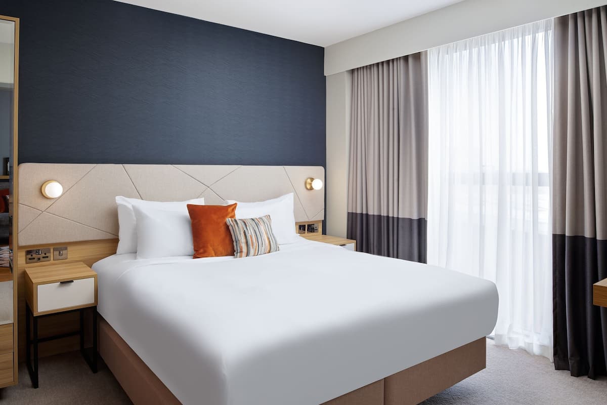 A hotel room with a large bed, two bedside tables, a nightstand, and a window with curtains.