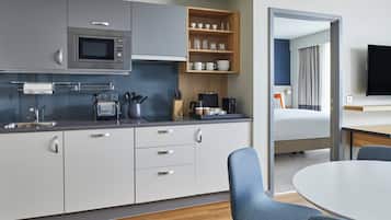 Una cocina moderna con armarios grises, un microondas, un tostador y una cafetera. Hay una estantería de madera con tazas y vasos, y un comedor con una mesa y sillas.