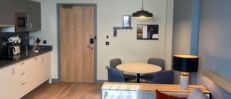 A modern kitchen with a microwave, toaster, and sink. There is a wooden door, a round table with chairs, and a sofa with a lamp.