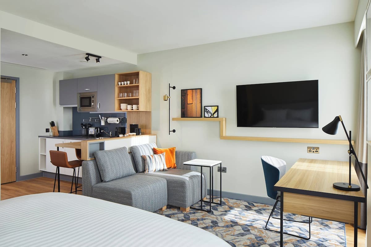A modern hotel room with a grey sofa, a small wooden desk, a TV mounted on the wall, and a kitchenette in the background.