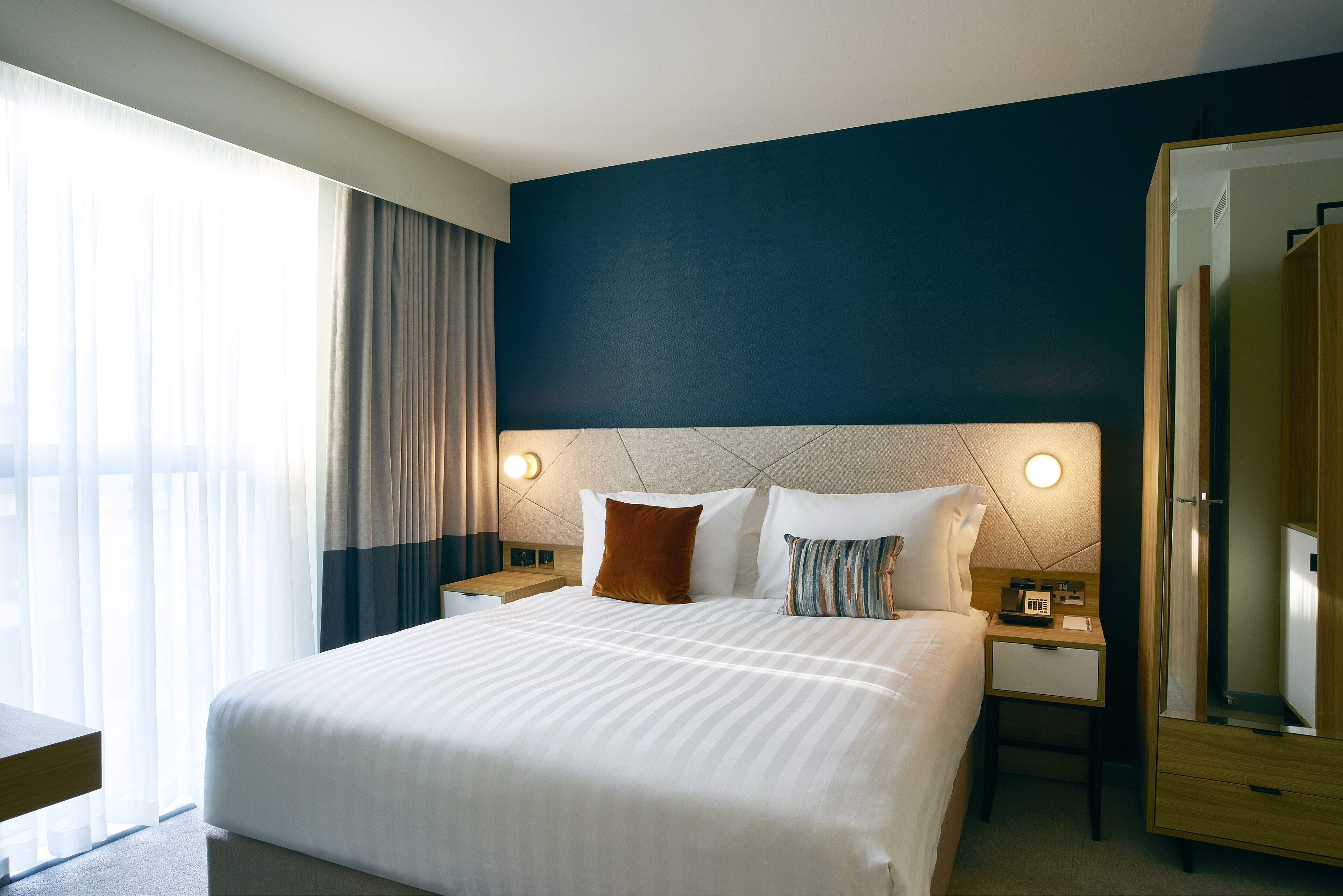 Habitación de hotel con una cama grande, mesitas de noche, un espejo y una ventana con cortinas.
