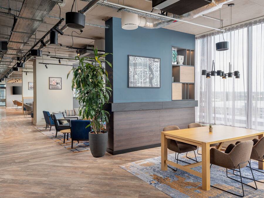 Un espacio de oficina moderno con una mesa de madera, sillas, una planta en maceta y un amplio ventanal con cortinas.