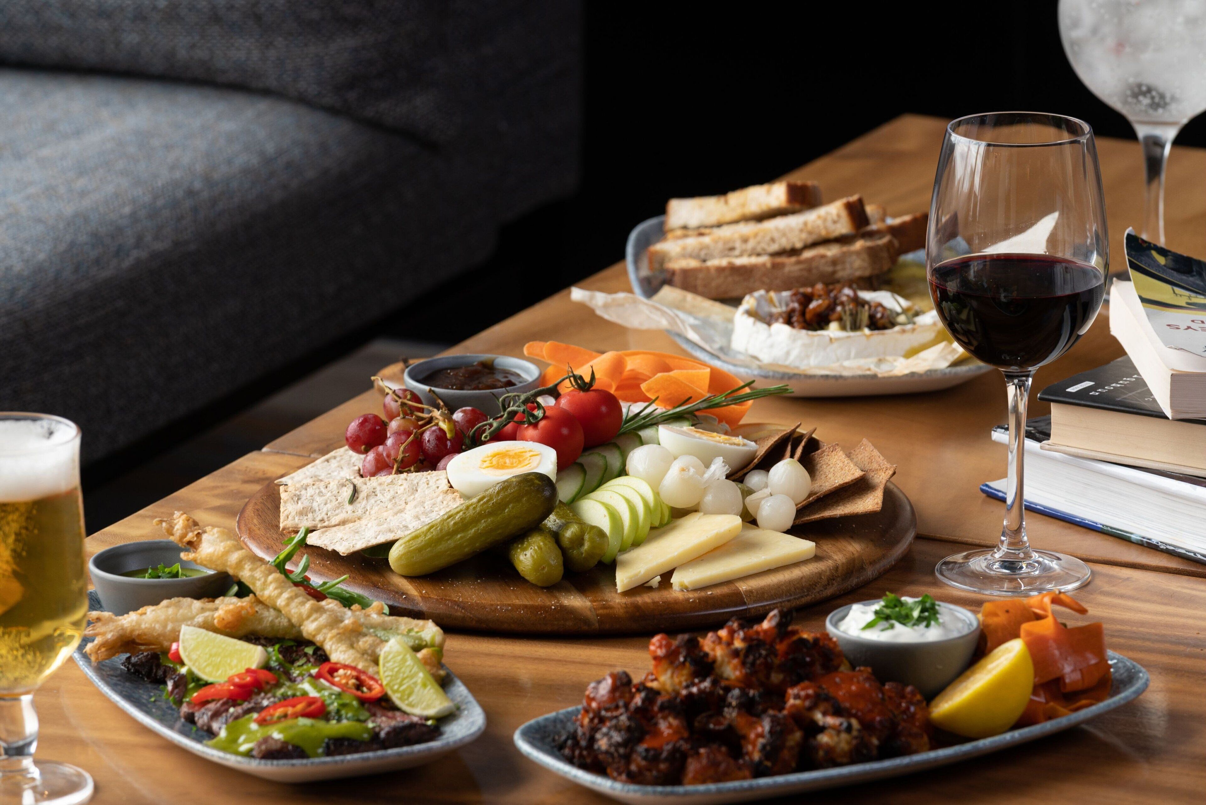 A wooden table with a variety of foods including bread, cheese, and vegetables, and a glass of red wine.