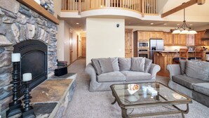 A spacious living area with a stone fireplace, a wooden balcony, and a kitchen with modern appliances. - Luxury Bigfork Retreat | Pond Views, Stone Fireplace & Steps from Flathead Lake | 30-Day Minimum (Bigfork)