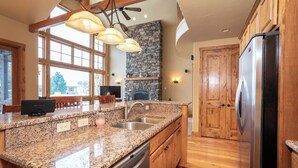 A kitchen with granite countertops, a stainless steel refrigerator, wooden cabinets, a stone fireplace, and a large window. - Luxury Bigfork Retreat | Pond Views, Stone Fireplace & Steps from Flathead Lake | 30-Day Minimum (Bigfork)
