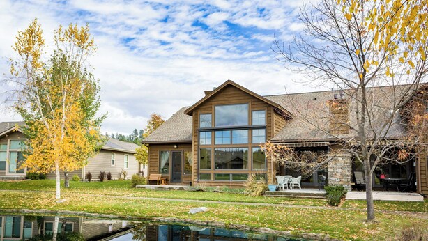 A modern house with a large window, a pond, and autumn trees. - Luxury Bigfork Retreat | Pond Views, Stone Fireplace & Steps from Flathead Lake | 30-Day Minimum (Bigfork)