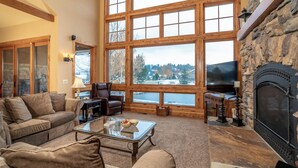 A spacious living room with a large stone fireplace, a stone wall, and a large window overlooking a snowy landscape. - Luxury Bigfork Retreat | Pond Views, Stone Fireplace & Steps from Flathead Lake | 30-Day Minimum (Bigfork)