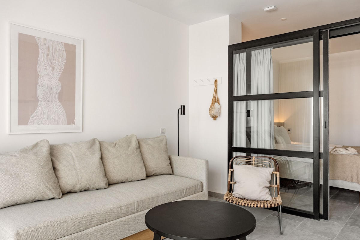 Una sala de estar moderna con un sofá, una mesa redonda y una silla de mimbre. Hay un ventanal con cortinas, una obra de arte enmarcada en la pared y una puerta corrediza de vidrio que da a otra habitación.