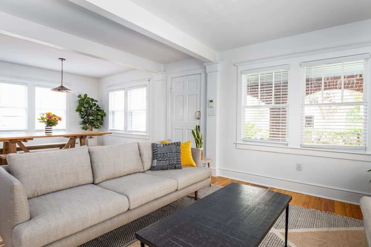 A living room with a sofa, a coffee table, and a dining table with chairs. There are windows with blinds, a door, and a ceiling light fixture.