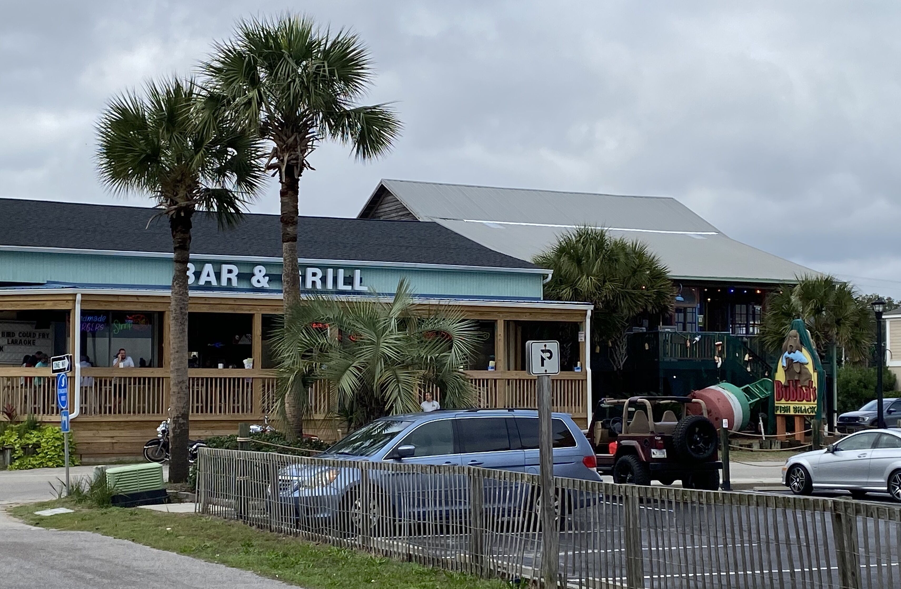 A restaurant with a bar and grill sign, outdoor seating, palm trees, and a parking lot with cars.