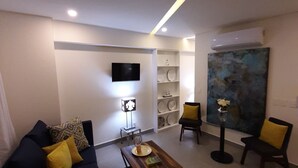 A modern living room with a sofa, coffee table, and chairs, a flat-screen TV, a painting, and a wall-mounted shelf.