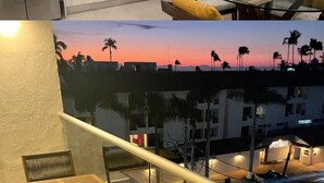 A modern kitchen with white cabinets, a stainless steel refrigerator, and a microwave. There is a dining area with a table set for two, yellow cushions, and a view of palm trees at sunset.