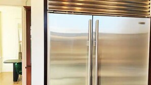 A stainless steel refrigerator with a double door design, located in a kitchen area with wooden flooring and a green chair visible in the background.
