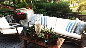 A wooden deck with a white sofa, a wicker chair, and a table with potted plants.