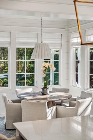 Una habitación con una mesa redonda, sillas, una lámpara colgante y ventanas amplias que dan a zonas verdes.