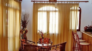 A dining area with a round table surrounded by wicker chairs, a vase with flowers, and a wooden bar counter with stools. - Anatoli Apartments (Hersonissos)