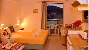 A hotel room with a bed, a small table with chairs, a window with a view of the city, and a kitchenette. - Anatoli Apartments (Hersonissos)