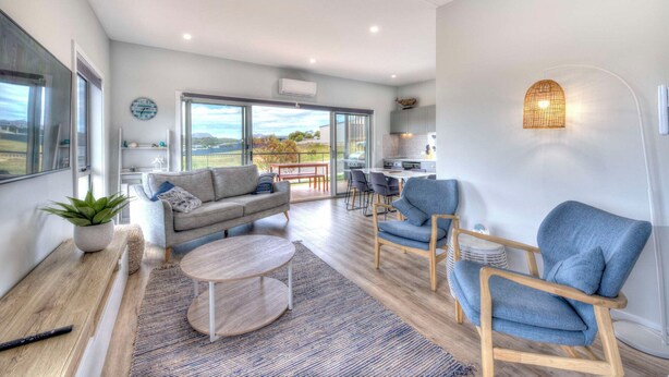 A spacious living room with a large window, a sofa, a coffee table, and a dining area with chairs. - Archella 2 - Freycinet Holiday Houses (Coles Bay)