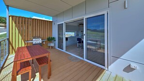 Eine Terrasse mit Holztisch und -stühlen, einem Grill und einer Glasschiebetür, die zu einem Küchenbereich mit Landschaftsblick führt.