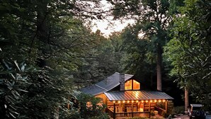 Une cabane rustique avec un toit en métal, entourée d’une forêt dense. Elle est dotée d’un porche décoré de guirlandes lumineuses et d’une fondation en pierre.