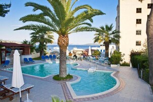 Un área de piscina con sillas de descanso y una palmera, rodeada por un edificio con numerosas ventanas.