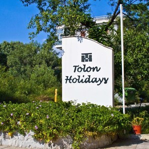 A sign for 'Tolon Holidays' with a logo, surrounded by greenery and a stone wall. - Tolon Holidays (Nafplio)