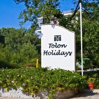 A sign for 'Tolon Holidays' with a logo, surrounded by greenery and a stone wall.