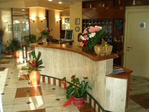 A hotel reception area with a marble counter, potted plants, and a sign indicating an exchange service. - Tolon Holidays (Nafplio)
