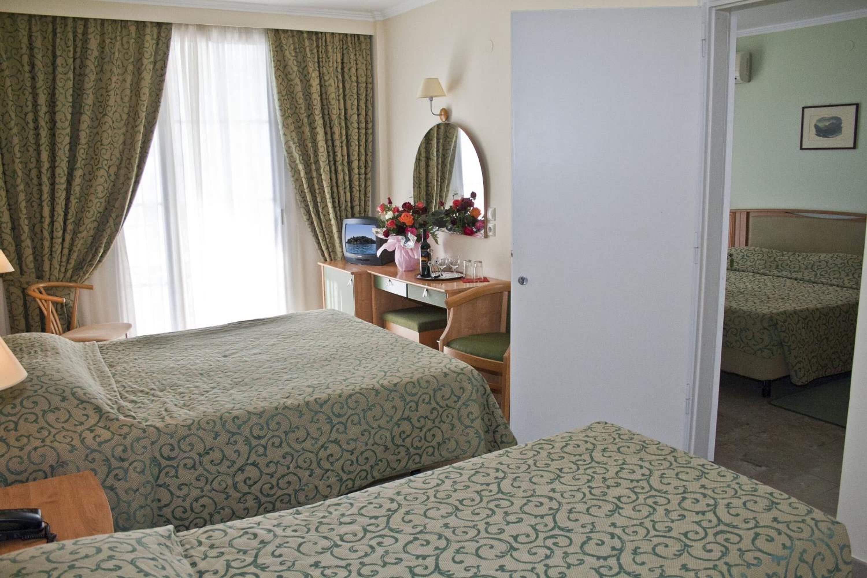A hotel room with two beds, a desk with a computer and flowers, a mirror, and a window with curtains.