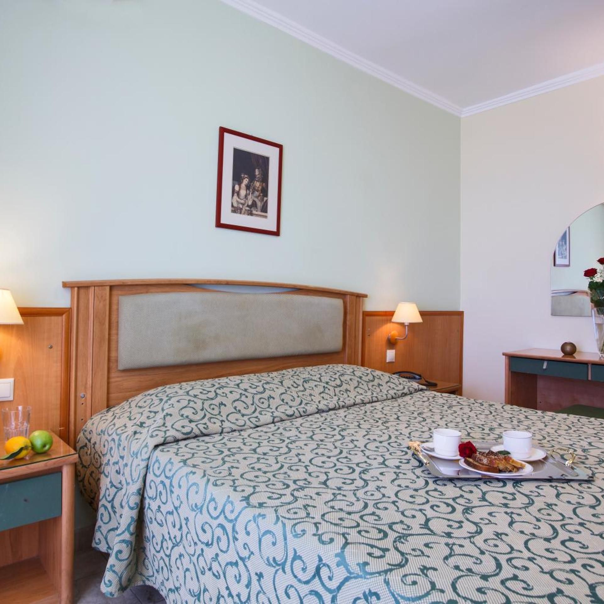 A hotel room with a bed, bedside tables, a lamp, a framed picture on the wall, and a tray with food and drinks.