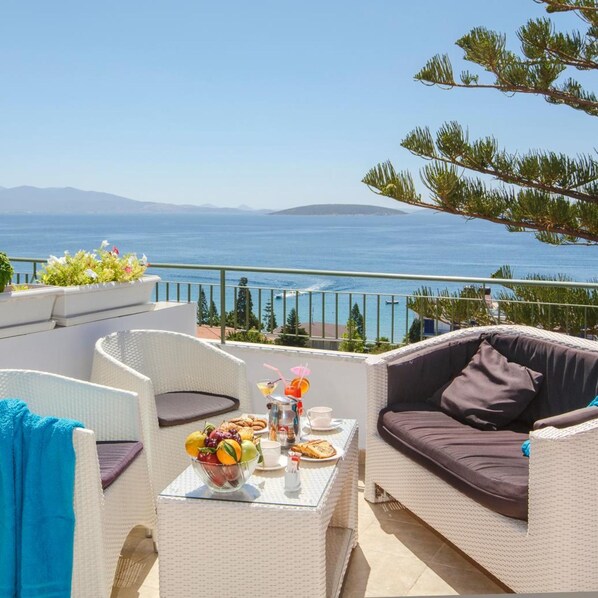 Una terraza con mobiliario de mimbre, una mesa con frutas y bebidas, y vistas al mar y a las montañas.
