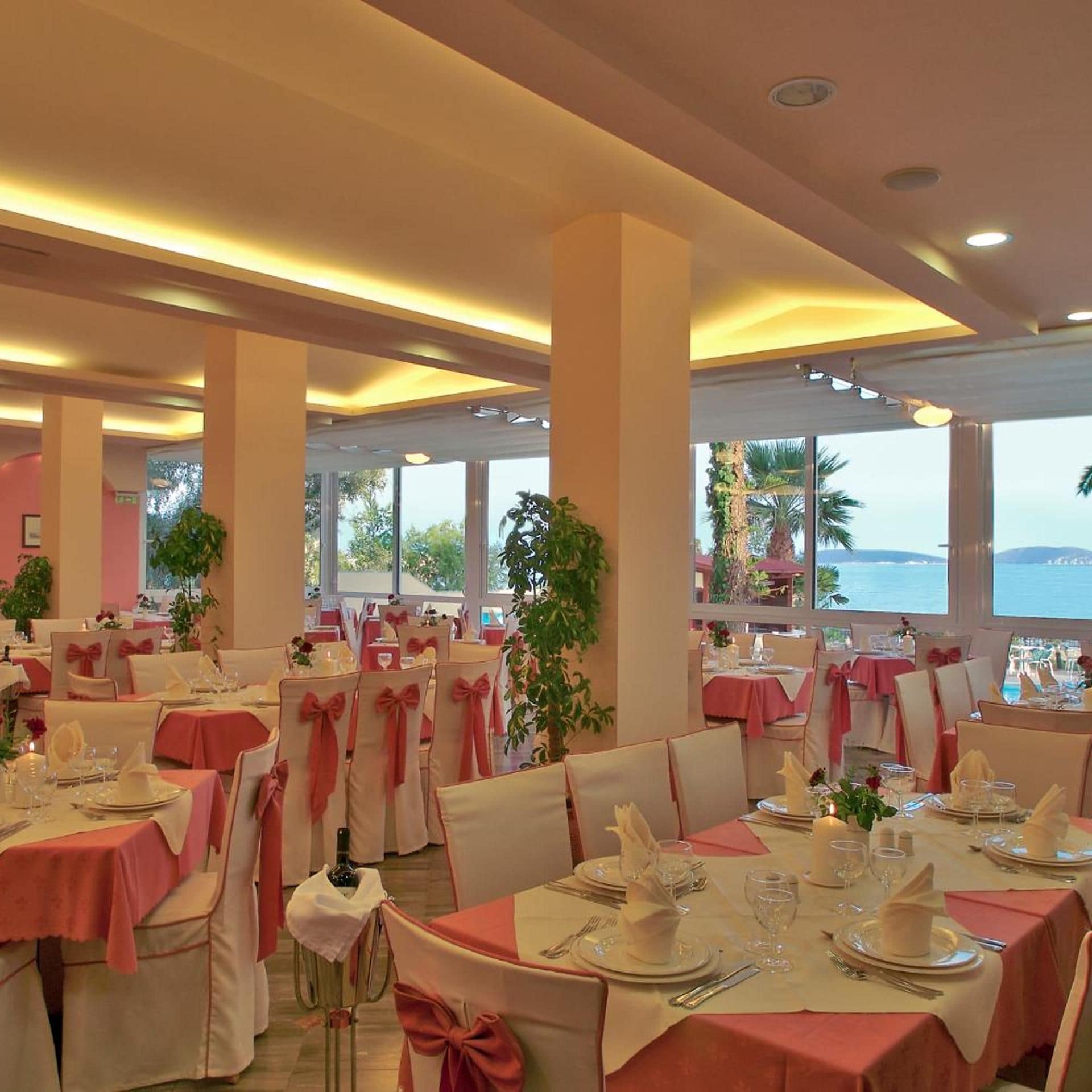 A restaurant with round tables, chairs with pink bows, and a view of the sea.